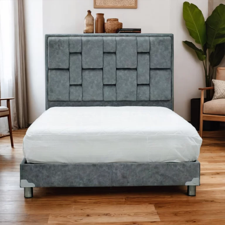 La imagen muestra un somier con un diseño moderno y elegante. El cabecero está tapizado en tela gris con paneles acolchados dispuestos en un patrón geométrico único, creando un efecto visual interesante. La base de la cama también está tapizada en tela gris y cuenta con patas metálicas que proporcionan soporte y estabilidad. La cama está vestida con una colcha blanca sencilla.