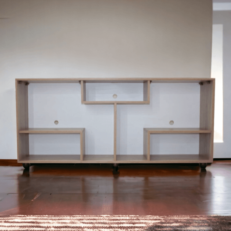 El centro de entretenimiento de la imagen presenta una estructura rectangular en madera clara, con un diseño moderno y asimétrico. Tiene varios compartimentos abiertos de diferentes tamaños distribuidos de manera irregular, creando un patrón visual interesante. El mueble está equipado con ruedas en la parte inferior para facilitar su movilidad. La parte trasera del mueble tiene orificios para el paso de cables, lo que lo hace ideal para la organización de dispositivos electrónicos.