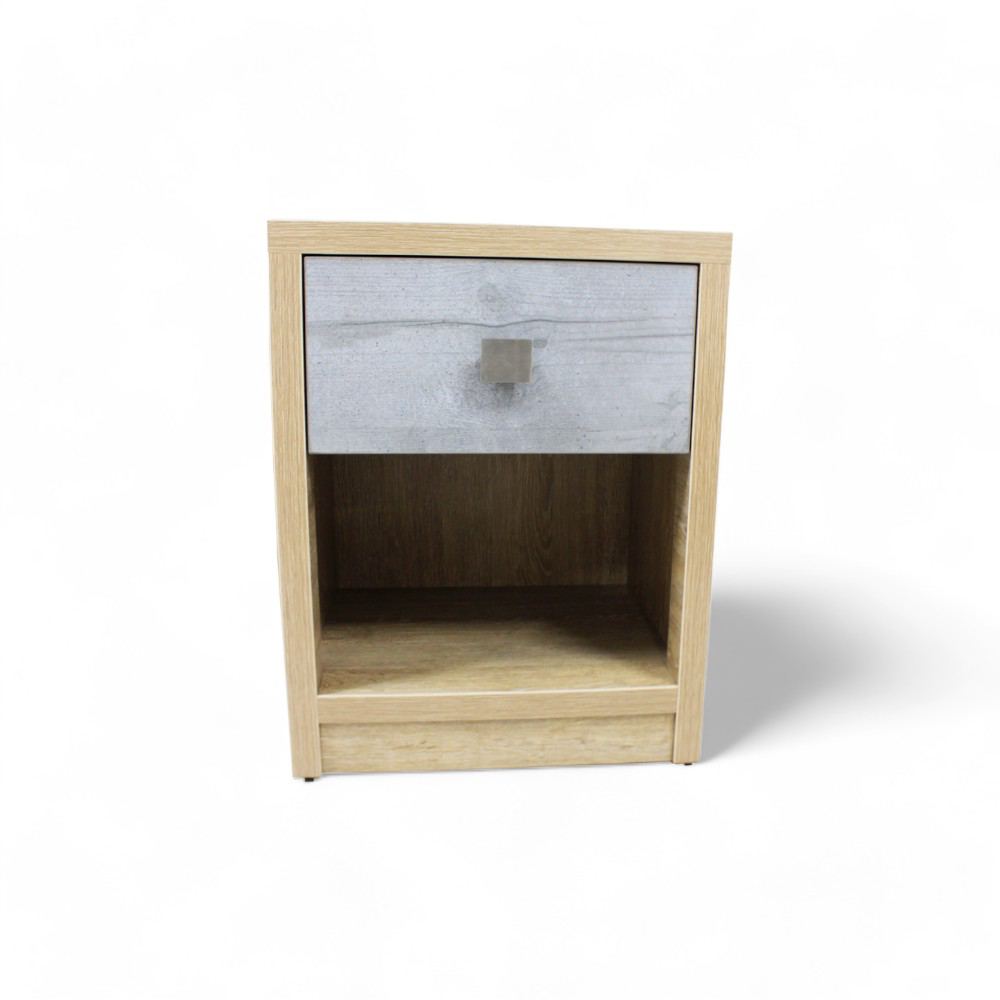 La imagen muestra una mesa de noche con un diseño simple y moderno. El buró está hecho de madera clara, y cuenta con un cajón en la parte superior. El frente del cajón tiene un acabado en color gris claro y una manija metálica cuadrada. Debajo del cajón, hay un espacio abierto que puede usarse para almacenamiento adicional.